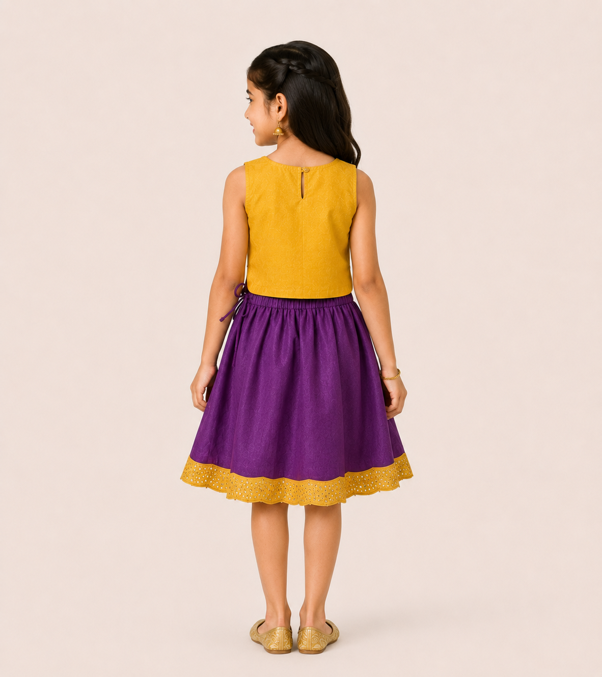 A young girl is shown from the back wearing a sleeveless two-tone dress with a mustard yellow top and a purple skirt with a yellow lace hem, set against a solid light beige background.
