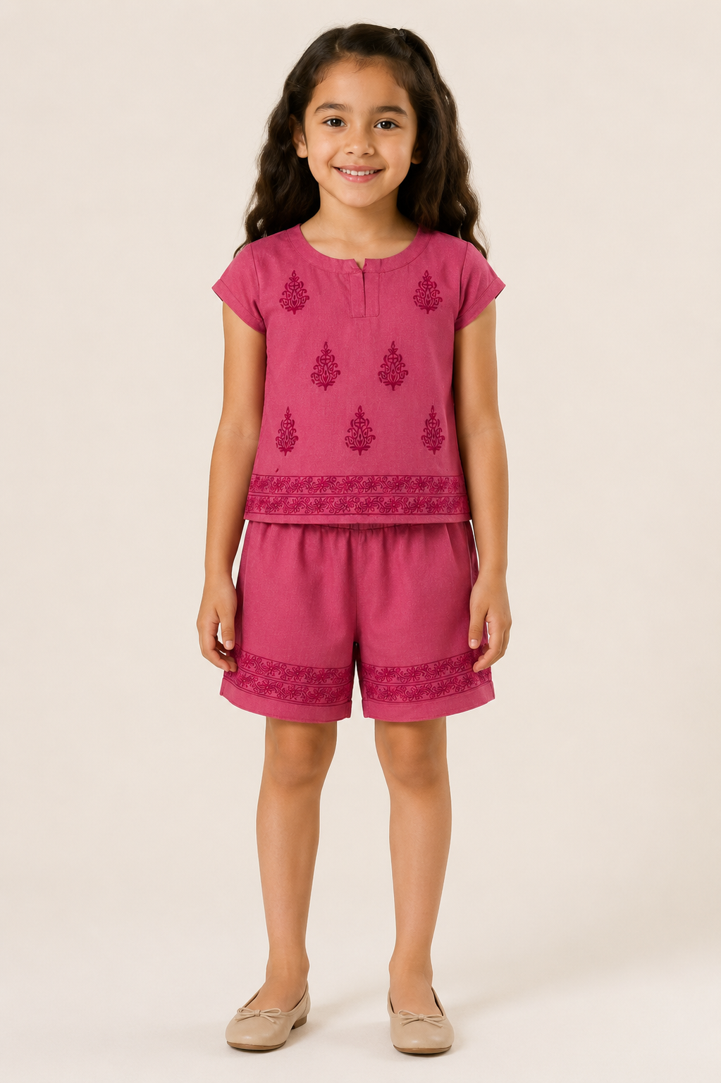 A young girl with long dark hair stands against a plain light beige studio background wearing a bright pink two-piece outfit consisting of a short-sleeve top and matching shorts, both with darker pink embroidered motifs and borders. She is wearing beige flat shoes.